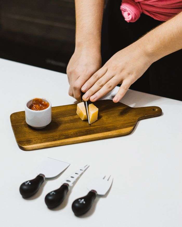 Hands cutting cheese on a wooden board with cheese knives and dip nearby, indoors.